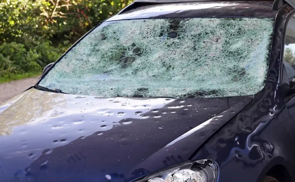 Car windshield severely damaged by hailstorm showing multiple cracks and shattered glass pattern across the front windshield of a parked vehicle.